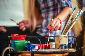 Middle part of girl artist paints portrait of woman with pencil. Girl painter seen from behind. Girl paints by pencil on easel. Paint brushes and foreground.