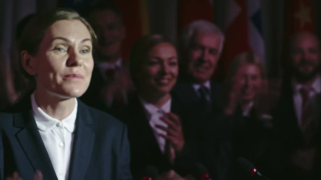 Tilt up shot of confident female politician speaking before public on conference with campaign members applauding in agreement