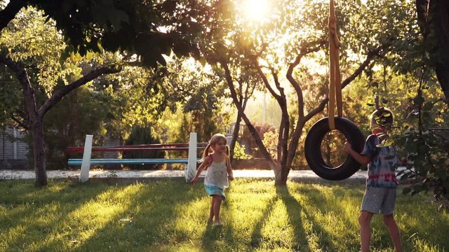 Slow motion of happy girl and boy playing at park with tire swing hanging from tree with beautiful sunlight in background