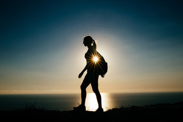 Fototapeta na wymiar silhouette Young woman tourist with backpack goes outdoor on the background on sunset sky and sea