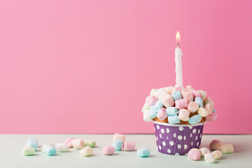 Delicious birthday vanilla cupcake with colorful marshmallows, cream and lighted candle on a pink...