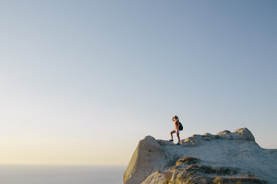 Young woman hiker with backpack standing on cliff and looking forward on the background of the sea, sky and city. lady tourist on top of a mountain enjoying view. Sport lifestyle travel concept