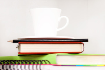Pencil on notebook near pile of books on shelf with cup of coffee