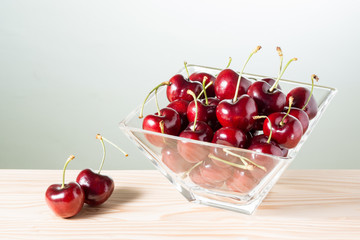 fresh red cherries in glass bowl and two fruit on pine wood table against bright background