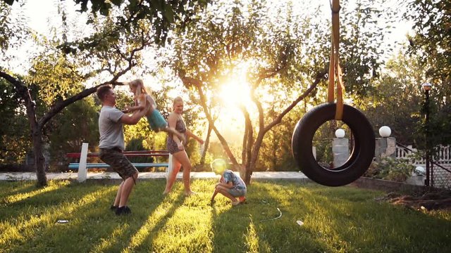 Parent playing with small two happy children at home on backyard with swing and trees during sunset. Slowmotion