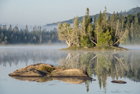partially submerged rocks in calm lake at sunrise, with lake mist and forest in background