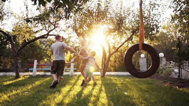 Children having fun holding hands with parents at park with tire swing in foreground and sunlight coming through trees. Having joined hands are turned around
