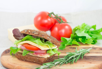 Sandwich with ham, tomato, cheese and lettuche on cutting board with rosemary on it. Closeup fast morning breakfast.
