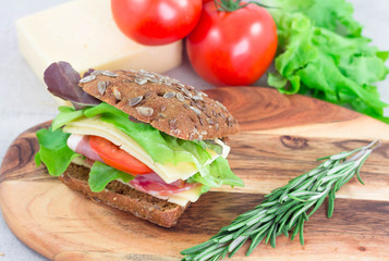 Sandwich with ham, tomato, cheese and lettuche on cutting board with rosemary on it. Closeup fast morning breakfast.