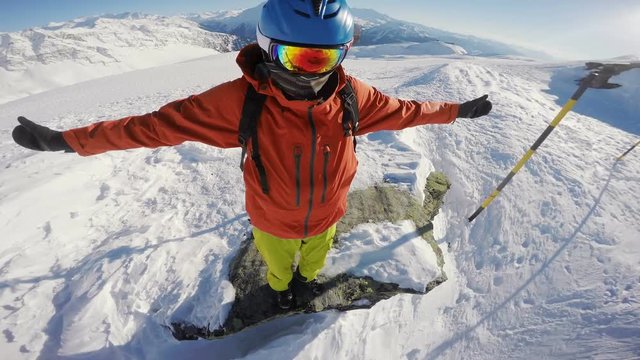 Snowboarder on top of the world, standing on the hill edge, looking down a mountain drop, arms outstretched, camera rotating around his head
