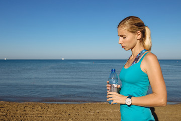 Portrait of young beautiful blonde woman wearing sportswear standing on the beach with a bottle of water after jogging or running..