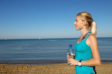 Portrait of young beautiful blonde woman wearing sportswear holding water from a bottle on background of the beach and sea.