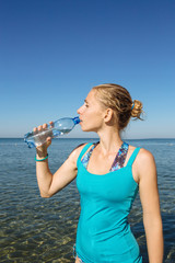 Portrait of young beautiful blonde woman wearing sportswear drinking water from a bottle after jogging or running on background of the  sea