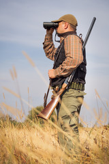 Man hunter with shotgun looking through binoculars in forest