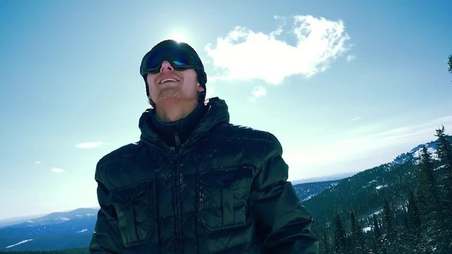 Portrait of smiling man in the blue skiing glasses against cloudy blue sky with the sun in slowmotion. 1920x1080