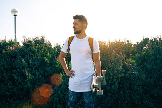 A scater with a skateboard in a blank white t-shirt is standing in a bushy area. A young man with a backpack is looking aside on a sunny day. Sun flare.