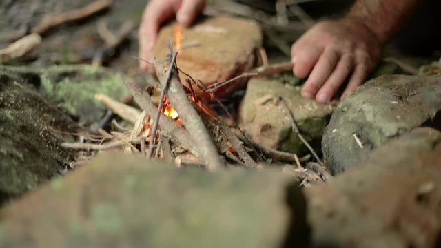 Handheld shot of a man building a small campfire using twigs in a fire ring.