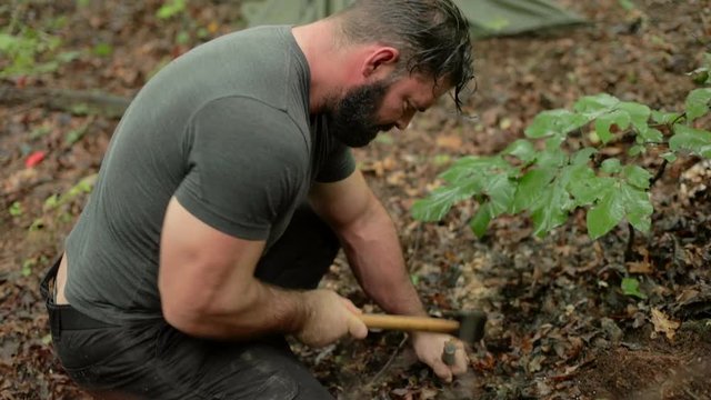 Handheld shot of a man hammering a surviver shelter stake into the ground using a hatchet.