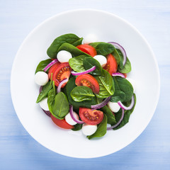 Fresh salad with mozzarella cheese, tomato, spinach and purple onion on blue wooden background top view. Healthy food.