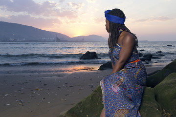 African woman in traditional dress, sitting on a stone at sunset, looking to the sea in a dirty beach.