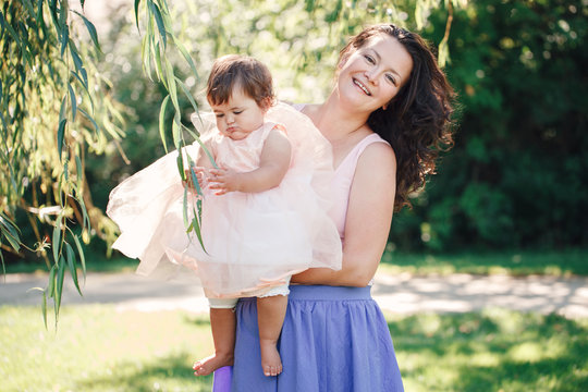 Lifestyle group portrait of smiling white Caucasian brunette mother holding hugging daughter in pink dress looking in camera on sunny spring summer day in park outside