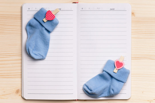 Baby Shoes And Notepad For Note On A Wooden Table. Blank Paper - View From Above.