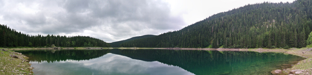 Panoramic image of lake landscape in the mountains, with pine trees reflecting on the water, on a cloudy day; nature with no people. Black lake, Durmitor, Montenegro. Horizontal panorama. - 120617081
