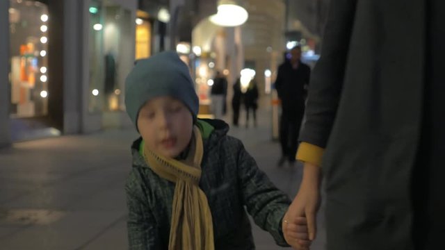 Little boy in overclothes holding mothers hand and talking to her while they walking in city street. Evening outing in pedestrian shopping area