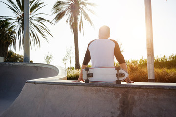 A male in a blank white long sleeve t-shirt is sitting with the back to the camera. Young man holding skateboard with the backside to the camera is sitting in the urban zone while relaxing. © JKstock