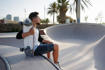 A brunet male in a blank white long sleeve t-shirt is holding the skateboard with the backside to the camera while having rest in skatepark. Young man is sitting in the urban zone while relaxing. © JKstock