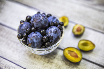 Слива и черноплодная рябина в вазе на деревянном столе. Рядом ягода и половинки сливы с косточками и без.