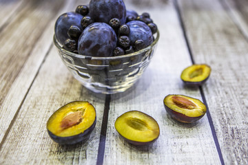 Слива и черноплодная рябина в вазе на деревянном столе. Рядом ягода и половинки сливы с косточками и без.