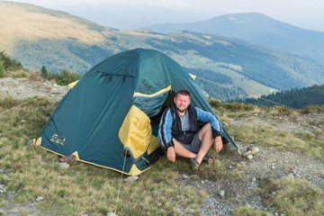 Tourist man sitting near a tent in the mountains looks at the camera