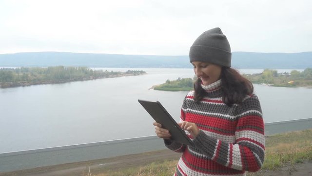 Young woman using tablet PC outdoors on background of the river and mountains. Autumn day.