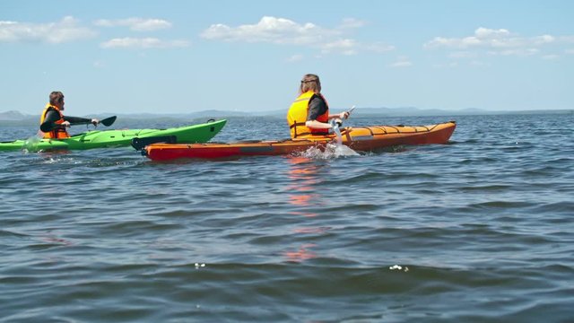 Slow motion rear view tracking of male and female athletes paddling with forward sweep strokes on kayaks along blue water surface on sunny day