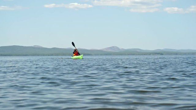 Slow motion lockdown of male kayaker paddling with forward sweep strokes along blue water lake in distance on sunny summer day