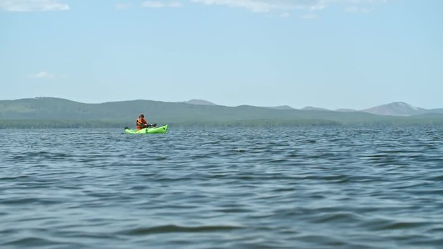 Slow motion of male kayaker paddling in distance with forward sweep strokes on blue lake water on sunny summer day, mountains visible in background 