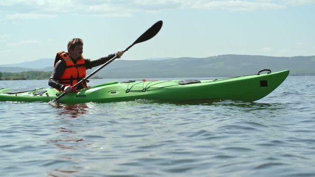 Tracking side view of male tourist skillfully paddling kayak on blue lake with misty mountains visible in distance