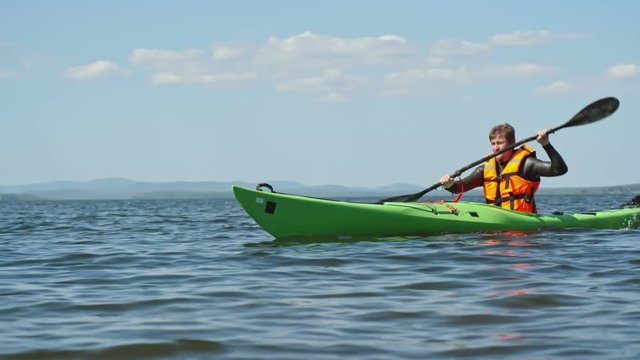 Slow motion tracking of male lake kayaker paddling with forward sweep strokes on blue water, misty mountains visible in distance 