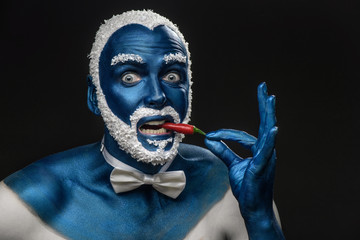 Man painted in blue color with snowy hair and beard eating chili pepper on black background