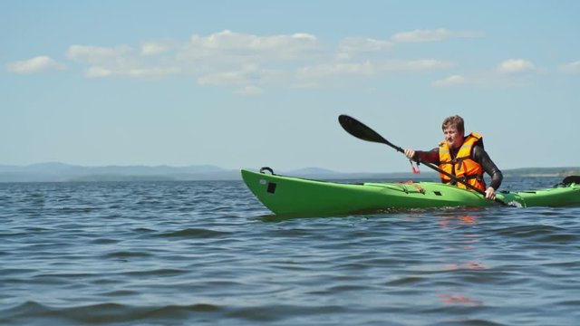 Slow motion tracking of middle aged tourist with beard paddling kayak on blue lake on sunny summer morning