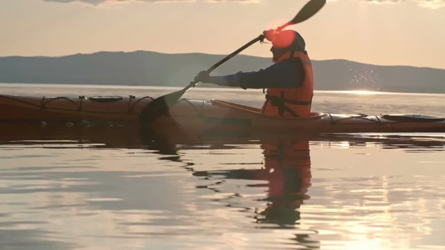 Slow motion tracking of man with beard paddling kayak on lake at sunset, chain of mountains visible in distance 
