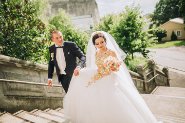 bride and groom walking near trees and church. Woman in yellow gold crown. Decorated yellow beads dress. Embracing couple. Looking each other. Adorable girl. Emotional picture.