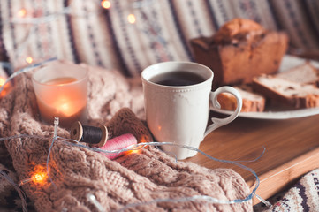 Cup of black tea with cake and candle staying on wooden tray in bed on knitted cloth closeup. Selective focus.