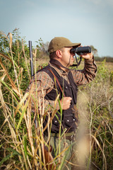 Man hunter with shotgun looking through binoculars in forest