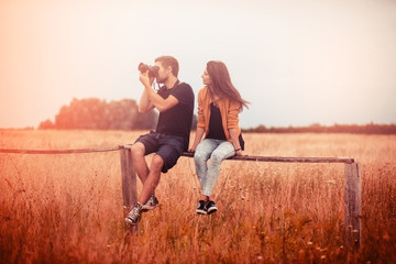 Young couple sitting on the fence in the summer sunlight and taking pictures with a digital camera, friendship and relationships concept