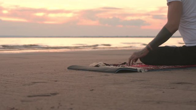 Young flexible tattoo man practicing yoga on the beach at sunset. Stretching exercises before practice.