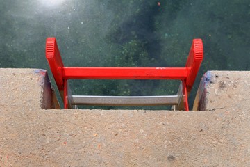 Bright red metal ladder on a concrete quayside leading down to the clear green water of a harbour, with the sun reflecting