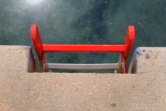 Bright red metal ladder on a concrete quayside leading down to the clear green water of a harbour, with the sun reflecting