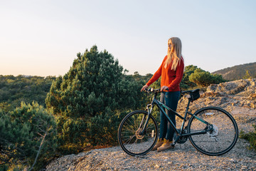 Fototapeta premium attractive young blonde woman in red sweater, blue jeans and yellow boots stands with bike on mountain road and looking forward